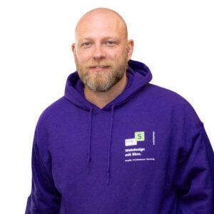 strg-S Webdesign mit Sinn Ein Mann mit rasiertem Kopf und Bart, der einen lila Kapuzenpullover mit der Aufschrift „Webdesign mit Sinn.“ trägt, steht vor einem schlichten weißen Hintergrund. WordPress, Shopify