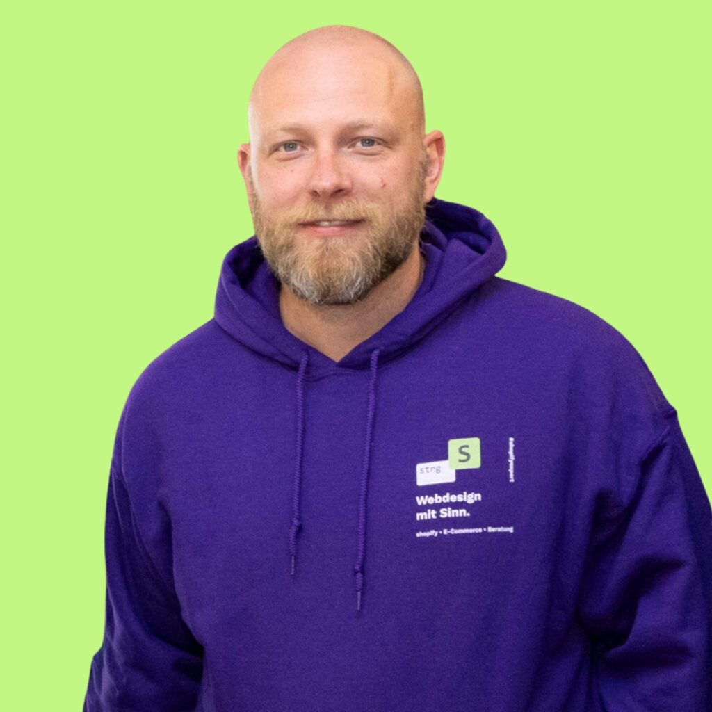 strg-S Webdesign mit Sinn Ein Mann mit Bart und lila Kapuzenpullover steht vor einem hellgrünen Hintergrund. Auf dem Kapuzenpullover ist weiß-grüner Text zu sehen. WordPress, Shopify